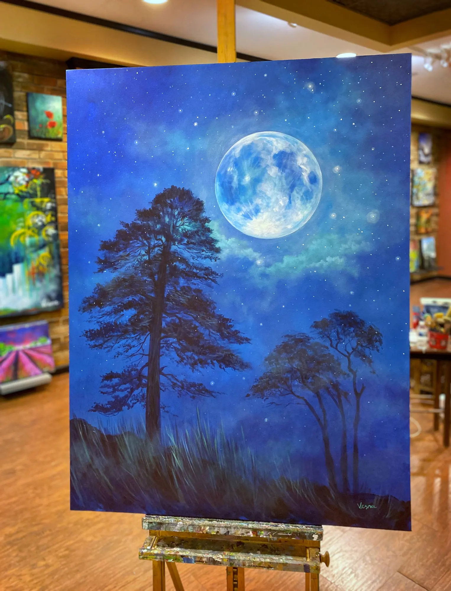 A night painting of pine trees under a full moon on an easel