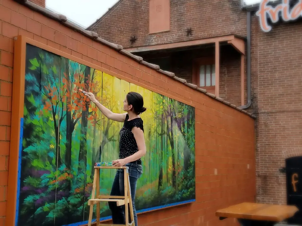 Vesna on a ladder painting a forest mural on an exterior wall